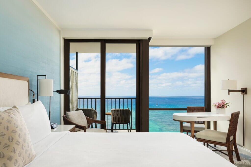 A bright hotel room with a neatly made bed, a small table with chairs, and a balcony overlooking the ocean under a partly cloudy sky.