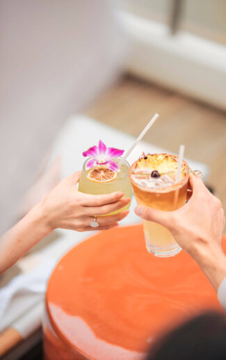 Two people toasting one another with tropical cocktails by the pool at Halepuna Hotel