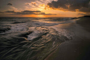 Waves gently crash onto a sandy beach under a dramatic sunset sky with dark clouds. The horizon glows orange and yellow, and two people stand near the water’s edge in the distance.