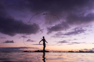 A silhouette of a person casting a fishing net stands in shallow water at sunset, with dramatic purple clouds in the sky and gentle water ripples in the foreground.