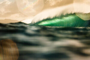 A large ocean wave curls and glows with green light under a cloudy sky, with sunlight creating lens flares and blurred foreground water.