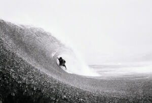 A surfer rides inside the barrel of a large, curling wave on a cloudy day, captured in a dramatic black and white photograph with water spraying around them.