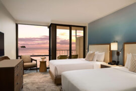 A modern hotel room with two double beds, a flat-screen TV, and a seating area by a sliding glass door. The glass door overlooks a balcony with a view of the ocean and a colorful sunset sky.