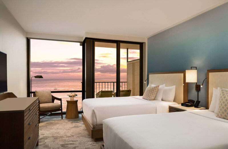 A modern hotel room with two double beds, a flat-screen TV, and a seating area by a sliding glass door. The glass door overlooks a balcony with a view of the ocean and a colorful sunset sky.