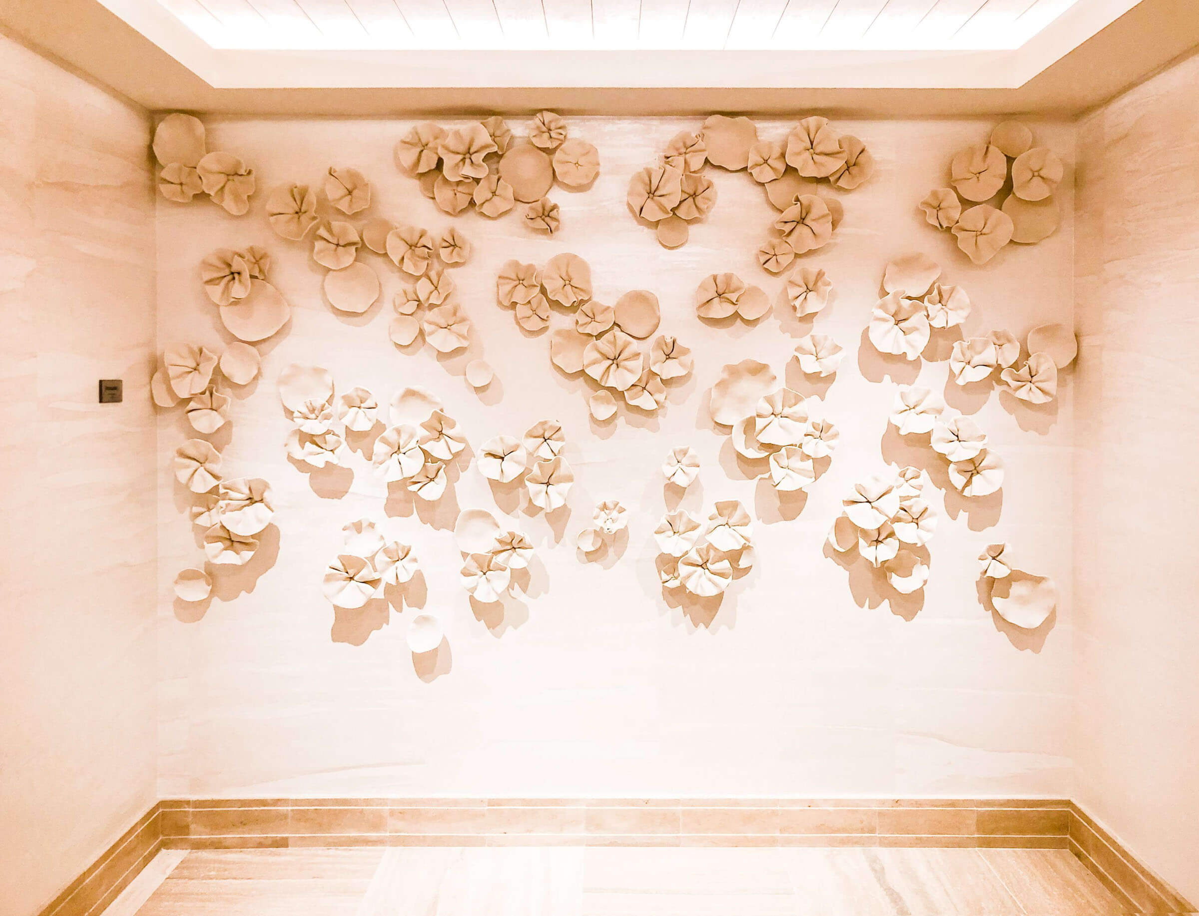 Wall art installation featuring clusters of cream-colored, abstract flower sculptures arranged on a light beige wall in a well-lit, modern hallway.