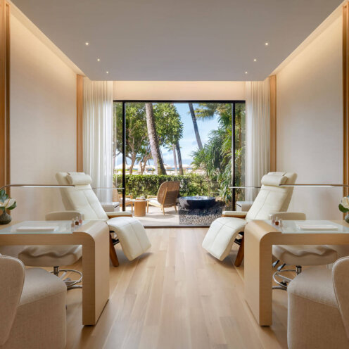 A bright, modern spa room with two manicure stations, plush cream chairs, light wood floors, and large windows overlooking palm trees and an outdoor seating area with a beach view.