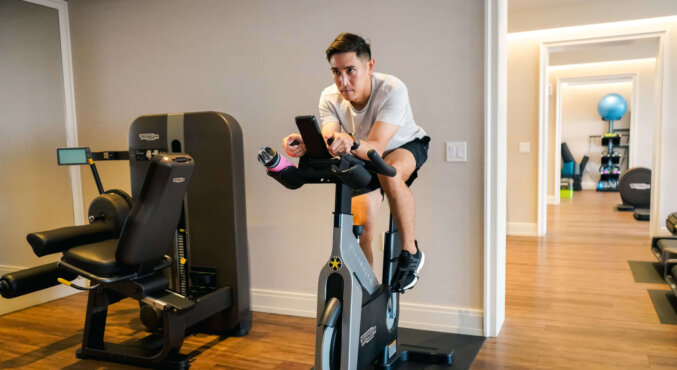 A man wearing a white shirt and black shorts rides a stationary exercise bike indoors, looking at his phone. There is gym equipment and wooden flooring in the room.