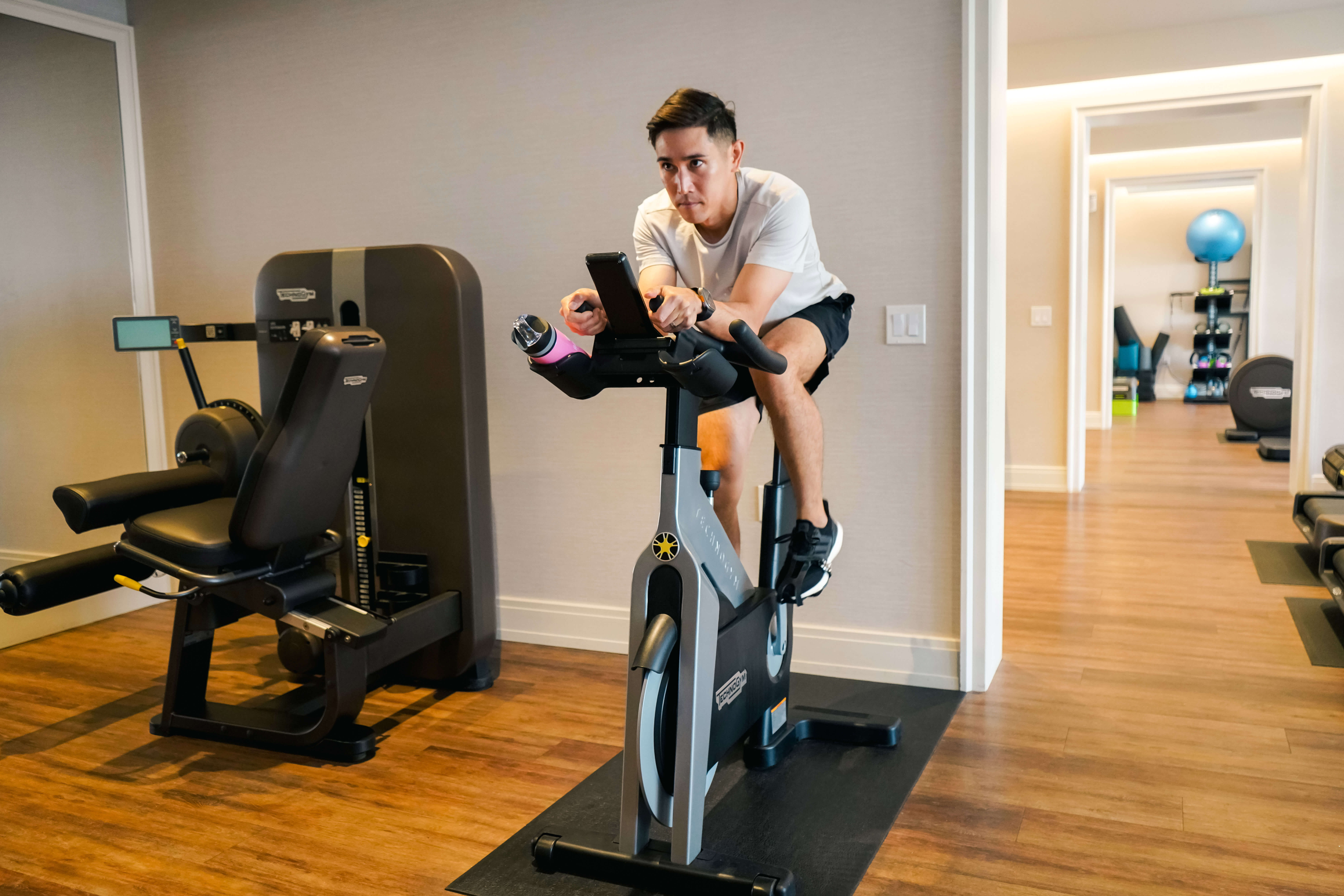 A man wearing a white shirt and black shorts rides a stationary exercise bike indoors, looking at his phone. There is gym equipment and wooden flooring in the room.