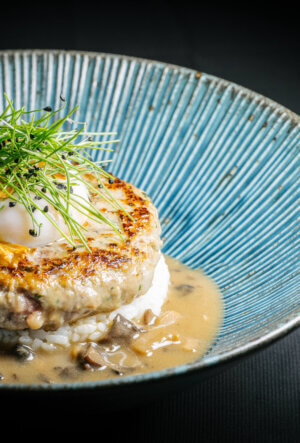 A close-up of a blue bowl with a hamburger steak on rice, topped with a soft-boiled egg and green garnish, surrounded by a creamy mushroom sauce.