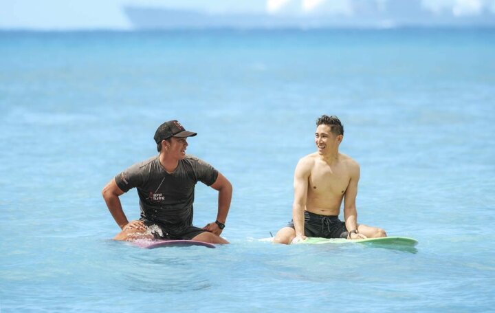 Two men sit on surfboards in the ocean, smiling and talking to each other. The sky and water are bright blue, and a blurry ship is visible in the background.