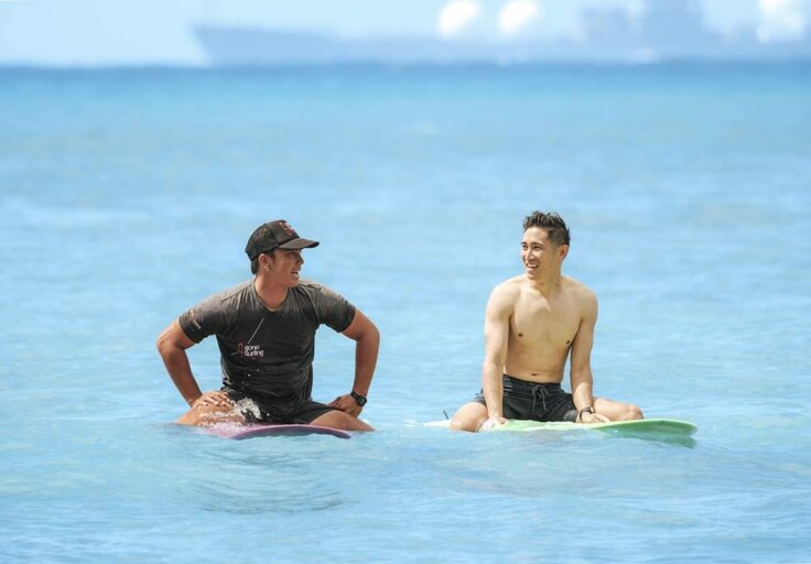 Two men sit on surfboards in the ocean, smiling and talking to each other. The sky and water are bright blue, and a blurry ship is visible in the background.
